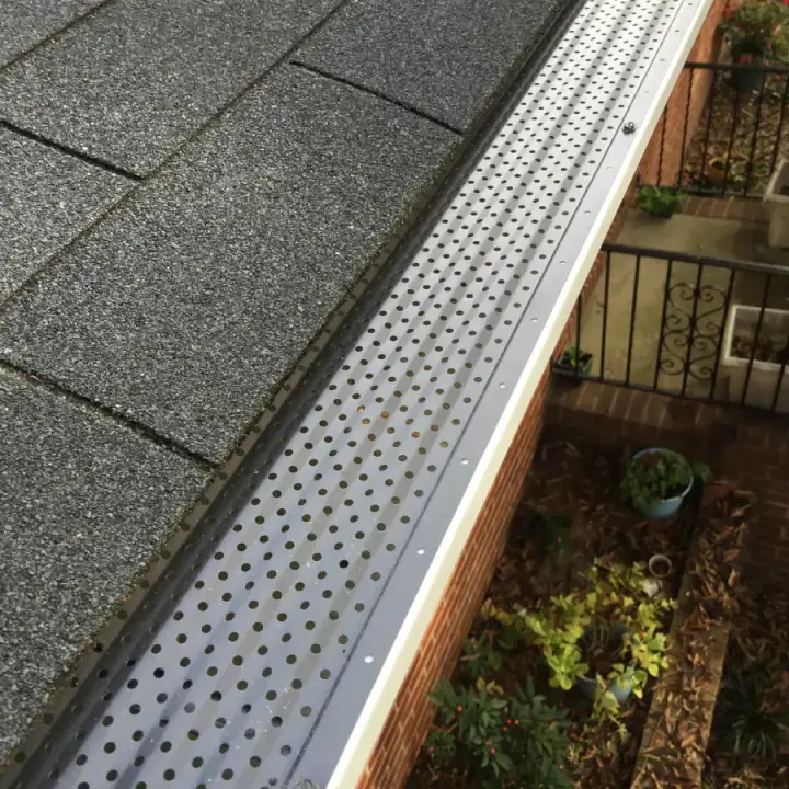 Perforated leaf guard fitted along a gutter below a shingle roof